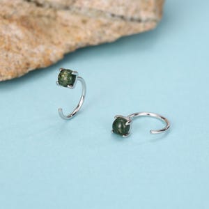 May include: Two silver-colored hoop earrings, each featuring a square-cut, dark green gemstone. The earrings are set against a light blue background, with a portion of a tan rock visible in the upper left corner.