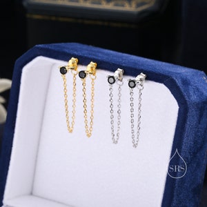 Pode incluir: Brincos de corrente dourados e prateados com detalhes de pedras preciosas pretas. Os brincos apresentam um pequeno pino com uma pedra preta, conectado a uma corrente delicada que pende por baixo. Os brincos são exibidos em uma caixa de joias de veludo azul.