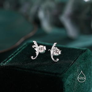 May include: A pair of silver earrings with a delicate, ornate design resembling a stylized leaf. The earrings are displayed on a green velvet box.