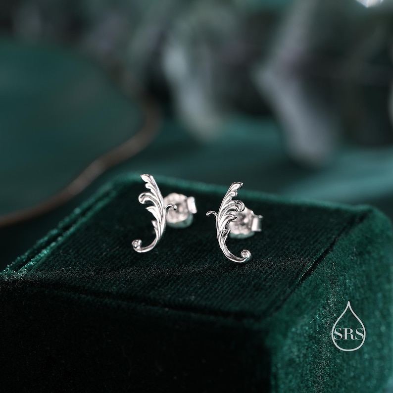May include: A pair of silver earrings with a delicate, ornate design resembling a stylized leaf. The earrings are displayed on a green velvet box.