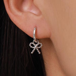 May include: A silver-colored earring with a bow-shaped charm. The earring is a small hoop with a twisted rope design on the bow. The earring is shown on a person's ear.