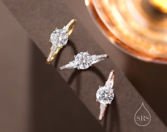 May include: Three engagement rings with different metal settings. The rings feature a round center stone with smaller accent stones on the band. The rings are displayed on a brown surface.