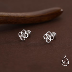 May include: A pair of silver stud earrings, each with a Celtic knot design. The earrings are shown on a brown surface, with a wooden element in the background. The earrings have a polished finish. The earrings are small and delicate.