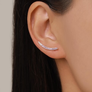May include: A silver ear climber earring featuring a curved design set with a row of small, vibrant pink gemstones. The piece follows the natural contour of the earlobe, creating a sleek and continuous line of colour against the skin.
