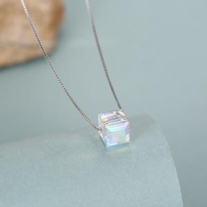 Pode incluir: Um colar de prata com um pingente de cubo transparente e iridescente. O pingente, com formato de cubo perfeito, reflete um arco-íris de cores. A delicada corrente de prata é exibida em um fundo azul claro.