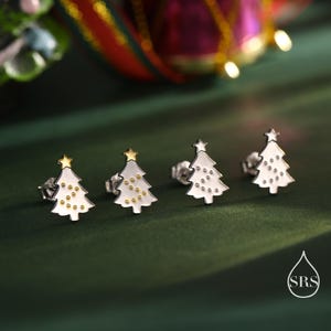 Könnte beinhalten: Vier silberne Weihnachtsbaum-Ohrstecker mit weißer Emaille und goldenen Tupfen. Jeder Baum ist mit einem goldenen Stern gekrönt. Die Ohrringe werden auf einer dunkelgrünen Oberfläche präsentiert.
