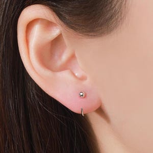 May include: Close-up of an ear with two silver earrings. One is a small ball stud, and the other is a small hoop earring. The skin is light-toned, and the earrings are shiny.