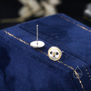 May include: A pair of silver owl stud earrings with gold accents. The owl's eyes are black and the beak is yellow. The earrings are on a blue background.