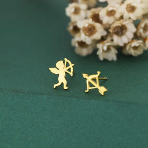 May include: A pair of gold-coloured stud earrings. One earring features a small angel with a bow and arrow, and the other earring is a bow and arrow. The earrings are on a dark green surface with white flowers in the background.