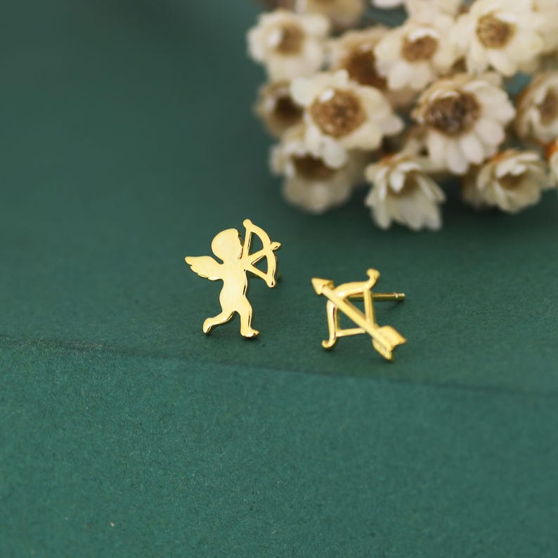 May include: A pair of gold-coloured stud earrings. One earring features a small angel with a bow and arrow, and the other earring is a bow and arrow. The earrings are on a dark green surface with white flowers in the background.