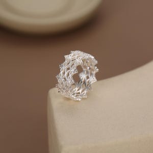 May include: A silver ring featuring an intricate, openwork lace-like pattern with a textured finish. The band has a scalloped edge design that creates a delicate, repeating geometric motif throughout the entire circumference of the metal ring.