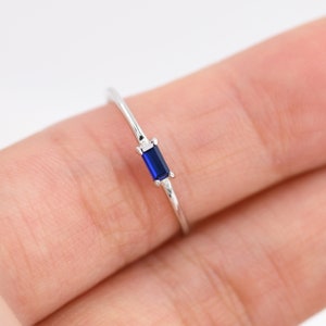 Könnte beinhalten: Ein zarter Silberring mit einem rechteckigen, tiefblauen Edelstein. Das schlanke Band ergänzt die lebendige Farbe des Steins und schafft ein minimalistisches und elegantes Design. Dieses Schmuckstück eignet sich perfekt für den Alltag oder besondere Anlässe.