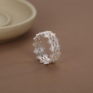 May include: A silver ring featuring an intricate, openwork lace-like pattern. The band is constructed from a delicate, repeating filigree design that creates a textured, scalloped edge along the top and bottom of the circular metal structure.