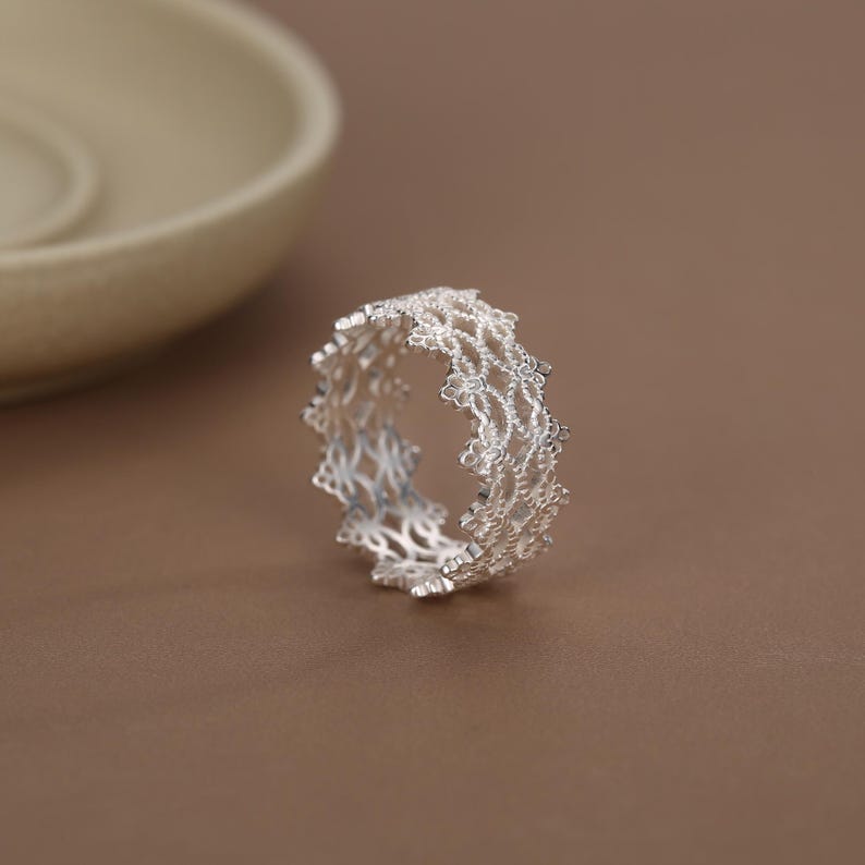 May include: A silver ring featuring an intricate, openwork lace-like pattern. The band is constructed from a delicate, repeating filigree design that creates a textured, scalloped edge along the top and bottom of the circular metal structure.
