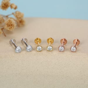 May include: Six stud earrings in silver, gold, and rose gold. Each earring has a round, clear gemstone in a bezel setting. The earrings are displayed on a light tan surface with a soft blue background.