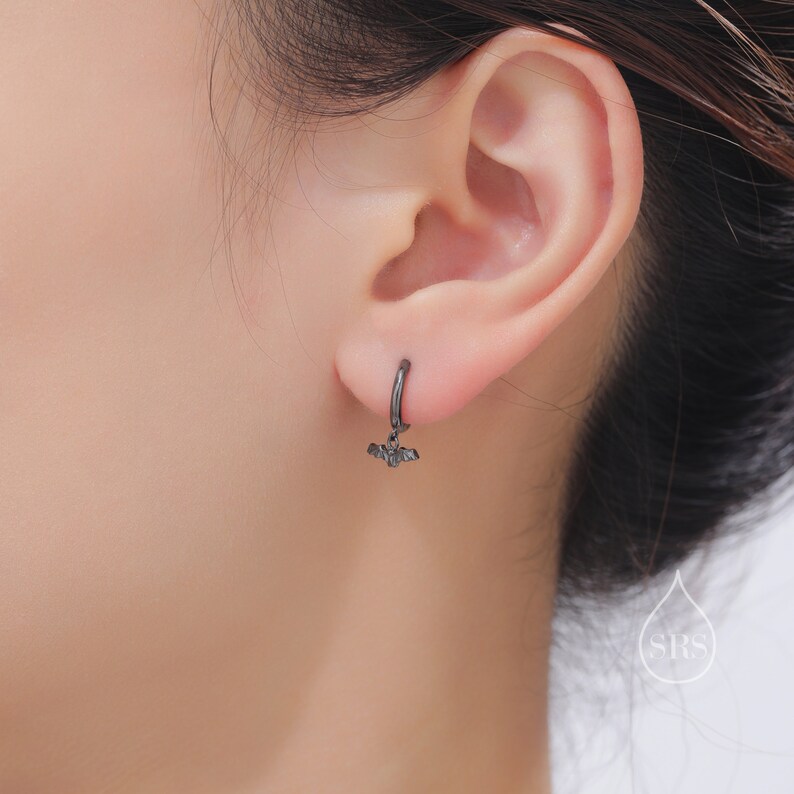 May include: A black hoop earring with a small bat charm hanging from it. The earring is worn in the left ear of a person.