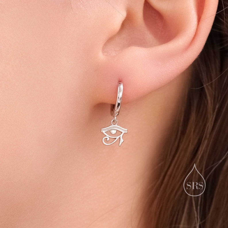 May include: Silver hoop earrings with a dangling charm of the Eye of Horus, a symbol of protection in ancient Egyptian mythology.