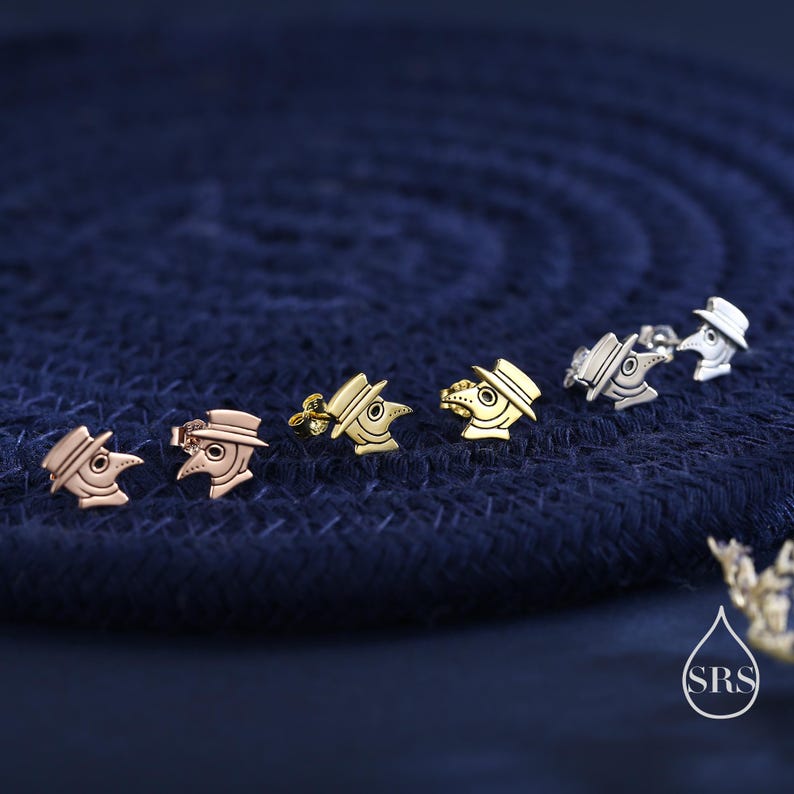 May include: A collection of stud earrings in various metallic finishes, including rose gold, gold, and silver. Each earring features a detailed design of a plague doctor's mask and hat, set against a textured navy blue background.