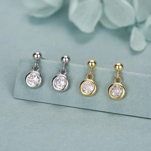 May include: Four pairs of stud earrings. Two pairs are silver-toned, and two are gold-toned. Each earring features a small, round, clear gemstone set in a bezel setting. The earrings are displayed on a light blue surface.