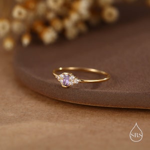 Puede incluir: Un delicado anillo de oro con una peque&ntilde;a piedra preciosa p&uacute;rpura y peque&ntilde;as piedras transparentes dispuestas en un racimo a su alrededor.