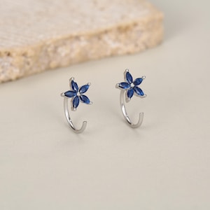 May include: A pair of silver-toned earrings featuring a flower-shaped design with five marquise-cut deep blue stones. Each earring has a curved, open-hoop style post that extends from the base of the floral arrangement, creating a delicate look.