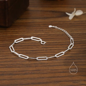 May include: A silver chain bracelet with a textured, rectangular link design. The bracelet is on a wooden surface.