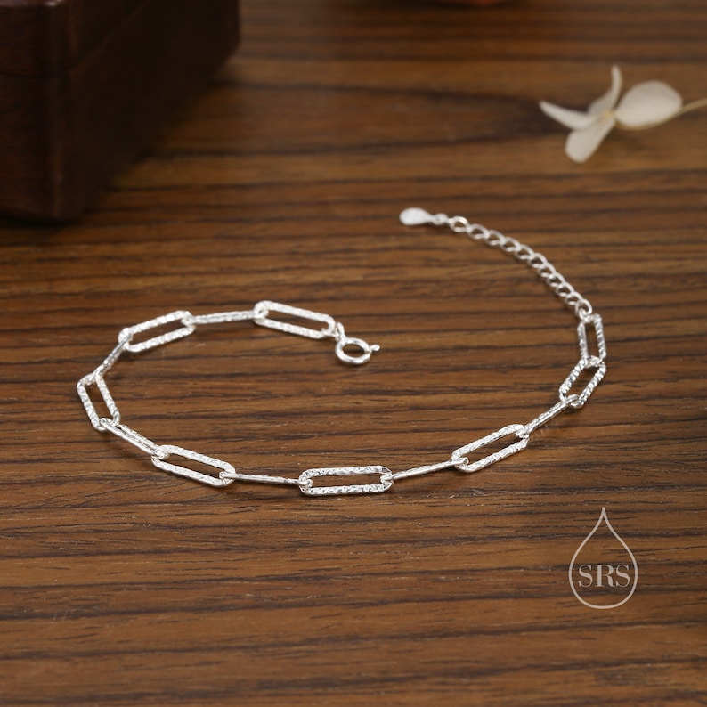 May include: A silver chain bracelet with a textured, rectangular link design. The bracelet is on a wooden surface.