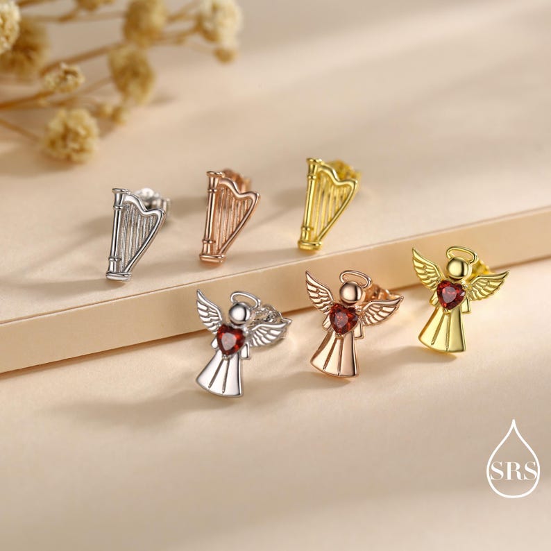 May include: A collection of stud earrings in silver, rose gold, and gold tones. The earrings feature harp and angel designs. The angel earrings have heart-shaped gemstones. The earrings are displayed on a neutral surface.