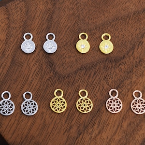 Op de afbeelding: Een verzameling kleine, decoratieve bedels in zilver, goud en roségoud. De bedels hebben verschillende ontwerpen, zoals een zonnestraal en een bloemmotief. Elke bedel heeft een kleine lus om aan sieraden te bevestigen.