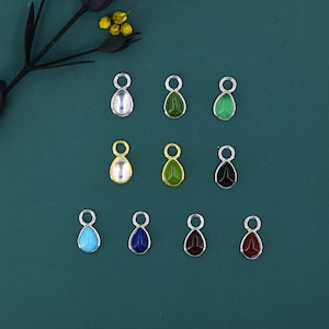 May include: Ten teardrop-shaped charms in a variety of colours, including silver, green, black, gold, blue, and red. Each charm has a small loop at the top for attaching to jewellery. The charms are arranged on a dark teal surface.