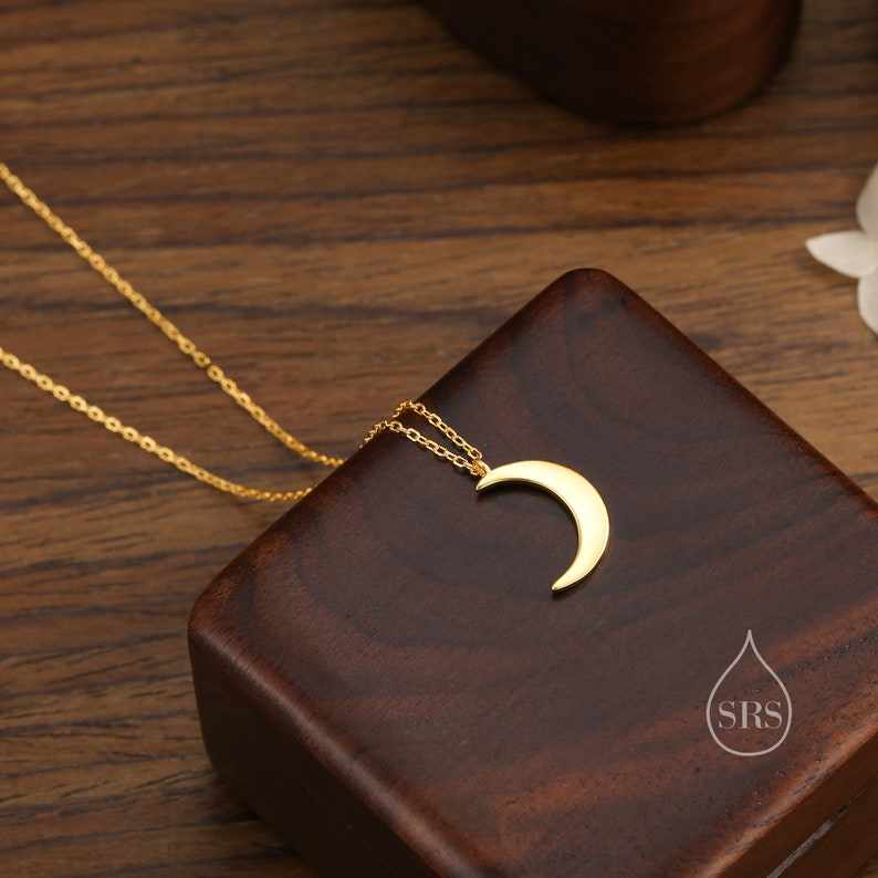 May include: A gold crescent moon pendant necklace on a delicate chain, displayed on a wooden box with the initials "SRS" in a water drop logo.