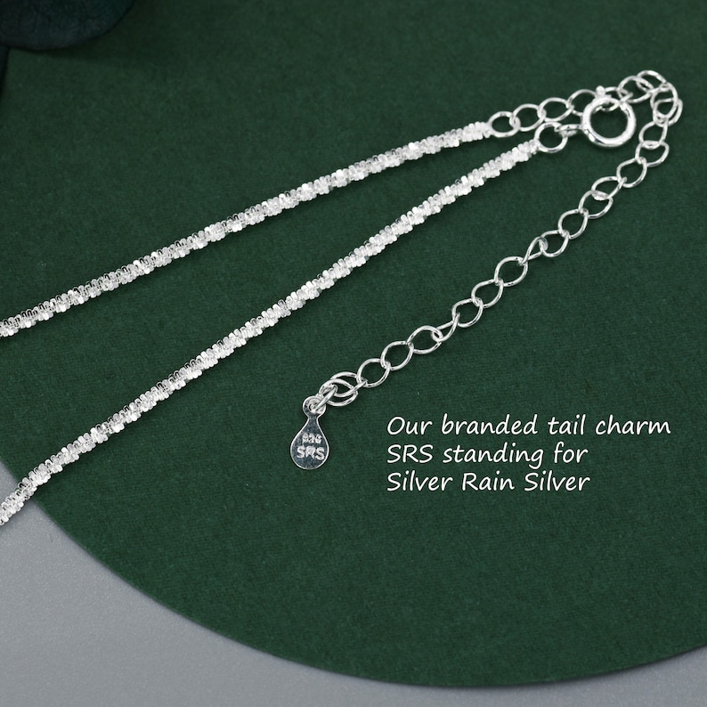 Puede incluir: Un collar de cadena de plata con un peque&ntilde;o dije de plata que dice "SRS". El dije est&aacute; unido al extremo de la cadena. La cadena est&aacute; sobre una superficie verde. Nuestro amuleto de cola de marca SRS significa Silver Rain Silver.