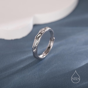 Puede incluir: Un anillo plateado con un diseño orgánico y texturizado. El anillo presenta una serie de cortes alargados en forma de óvalo a lo largo de su banda. El anillo está sobre un fondo azul, con una forma blanca y curva en el fondo.