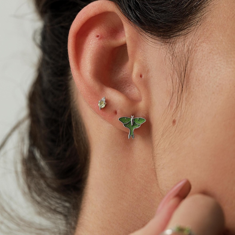 May include: A silver stud earring shaped like a green moth with detailed wings and a smaller, separate silver stud earring featuring a single, small, light green gemstone, both worn in the earlobe and upper ear cartilage respectively.
