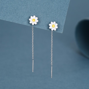 Può includere: Un paio di orecchini a forma di margherita con petali bianchi e centri gialli. Gli orecchini hanno lunghe e delicate catene d'argento che pendono dai perni dei fiori. Un accessorio a tema floreale.