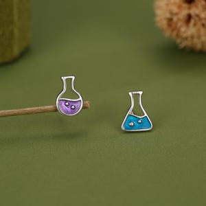 May include: A pair of stud earrings shaped like laboratory flasks. One earring features a purple enamel fill with two small silver bubbles, while the other earring features a bright blue enamel fill with two small silver bubbles, both with silver outlines.