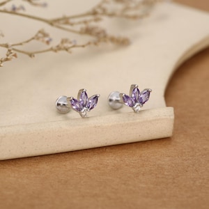 May include: A pair of silver stud earrings with a floral design. Each earring features three purple marquise-cut gemstones and a small white gemstone. The earrings are displayed on a neutral-coloured surface.