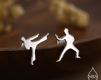 May include: A pair of silver-colored stud earrings. Each earring features a stylized silhouette of a person in a martial arts pose. One is kicking, and the other is in a defensive stance. The earrings are set against a blurred, warm-toned background.