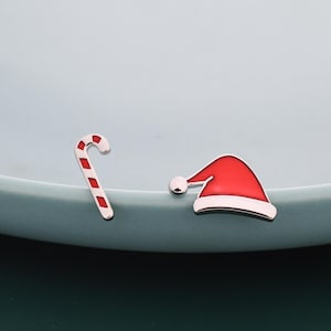 Puede incluir: Amuletos de esmalte con temática navideña. Un amuleto es un bastón de caramelo rojo y blanco, y el otro es un gorro de Papá Noel rojo con ribete blanco y pompón plateado. Los amuletos están sobre un fondo azul claro.