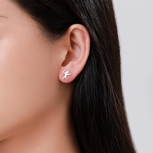 May include: A pair of silver stud earrings, each depicting a small Cupid figure with a bow and arrow. The earrings are displayed on an ear, showcasing their design. These earrings are a romantic accessory.