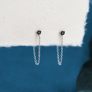Op de afbeelding: Een paar zilveren kettingoorbellen met zwarte edelstenen aan de bovenkant. De kettingen hangen naar beneden vanaf de edelstenen.