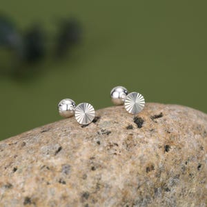 May include: A pair of silver stud earrings. Each earring has a small, round, polished silver ball and a flat, circular disc with a sunburst pattern. The earrings are displayed on a textured, light brown stone against a green backdrop.