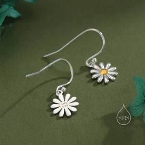 May include: A pair of silver dangle earrings with a daisy charm. The daisy charm has a yellow center and white petals. The back of the charm is stamped with "SRS 925".