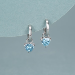 May include: Silver hoop earrings, each with a triangular, light blue gemstone pendant. The earrings are set against a light blue backdrop, highlighting the jewellery's design and colour.
