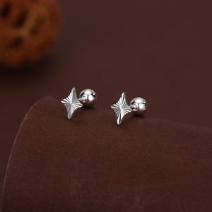 May include: A pair of silver star-shaped stud earrings. Each earring has a four-pointed star design with etched lines, paired with a small, round backing. The earrings are presented on a brown surface.