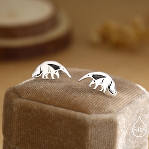 Pode incluir: Um par de brincos de prata com design de tamanduá. Cada brinco apresenta um tamanduá branco com detalhes em preto. Os brincos são exibidos em uma superfície de veludo marrom.