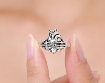 Puede incluir: Un anillo de tono plateado que presenta un diseño detallado de corazón anatómico sostenido por dos manos esqueléticas. El metal tiene un acabado oxidado para resaltar las texturas intrincadas de las cámaras cardíacas y la estructura ósea de los dedos.