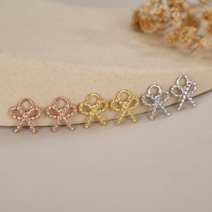 Op de afbeelding: Zes kleine strikvormige bedels in roségoud, goud en zilver. Elke bedel heeft een lusje voor bevestiging en een gedetailleerde touwachtige textuur. Ideaal voor het maken van sieraden of knutselen. Gepresenteerd op een neutrale achtergrond.