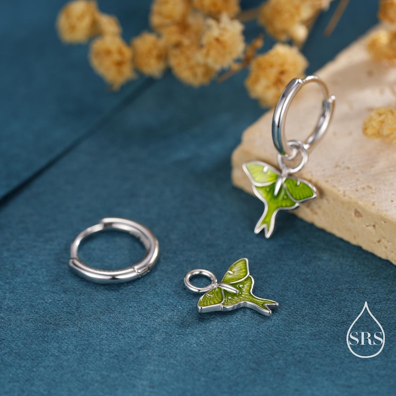 May include: Silver hoop earrings with a green and white moth charm. The earrings are displayed on a teal surface with dried flowers in the background. The earrings have a delicate design and are suitable for everyday wear.