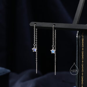 Op de afbeelding: Een paar zilveren oorhangers met een maansteenster-bedel aan elke oorring. De oorringen hangen aan een delicate ketting.